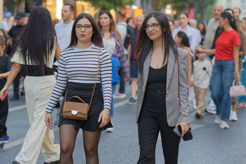 24 - Two women walking down a city street, with the woman on the left wearing a black-and-white striped long-sleeve top and a dark skirt, and the woman on the right wearing a black top